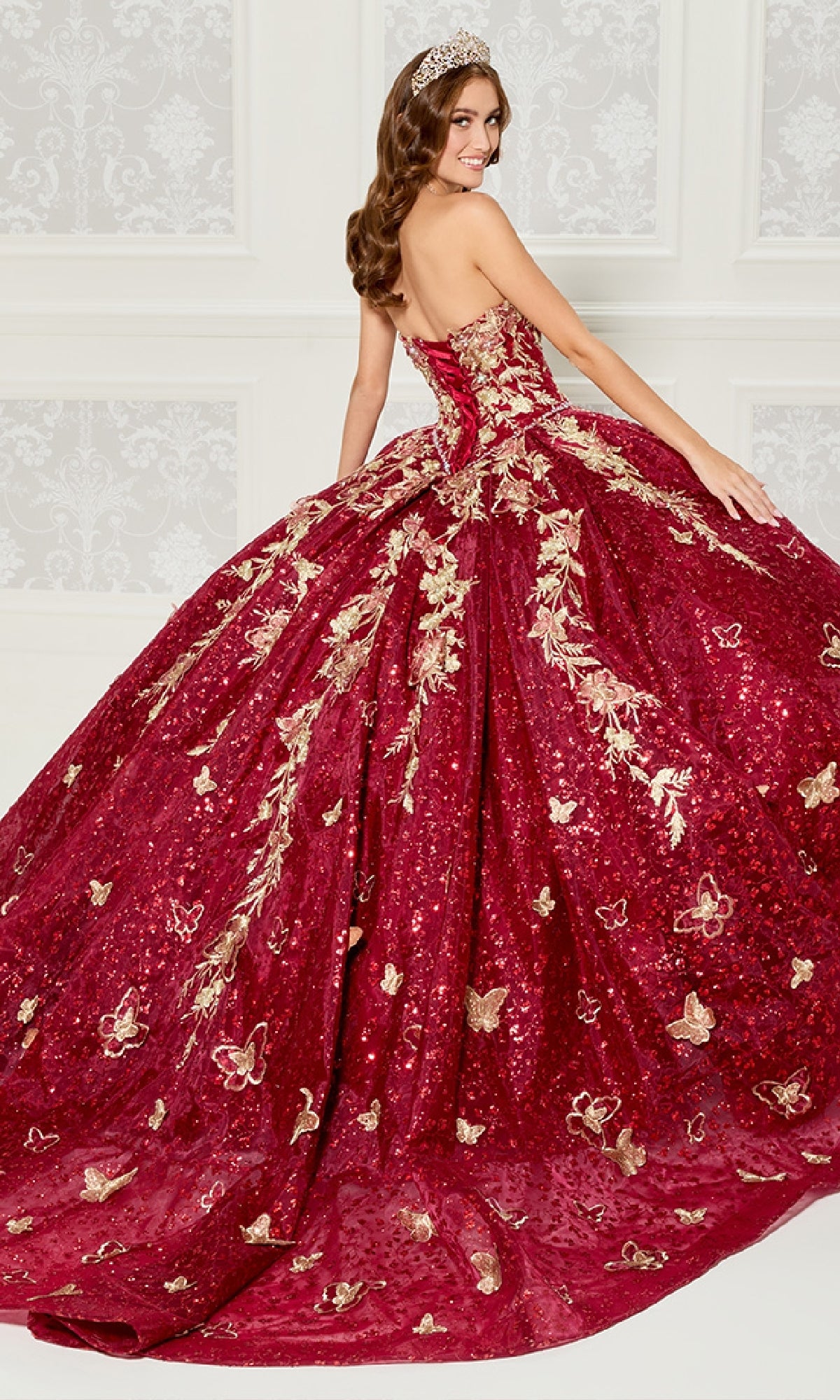 A woman stands gracefully in an elegant room, wearing the Strapless Quinceanera Dress: Princesa PR30121 in deep red with gold floral and butterfly embellishments, embroidered lace accents, and a tiara.